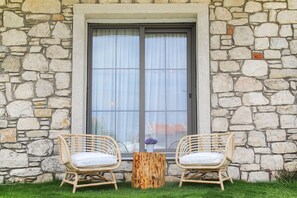 Two wicker chairs with white cushions and a wooden table with a vase of flowers in front of a stone wall with a large window. - Huzurla Otel (Urla)