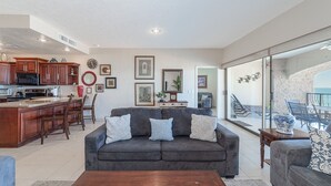 Een ruime woonkamer met een donkergrijze bank, een houten salontafel en een keuken met houten kastjes en een magnetron.