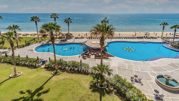 Un resort con una grande piscina centrale, piscine secondarie più piccole e una spiaggia attrezzata con ombrelloni.