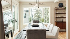 Uma área de refeições com uma mesa de madeira, um banco e um sofá branco. Há uma janela com vista para árvores e uma paisagem coberta de neve.