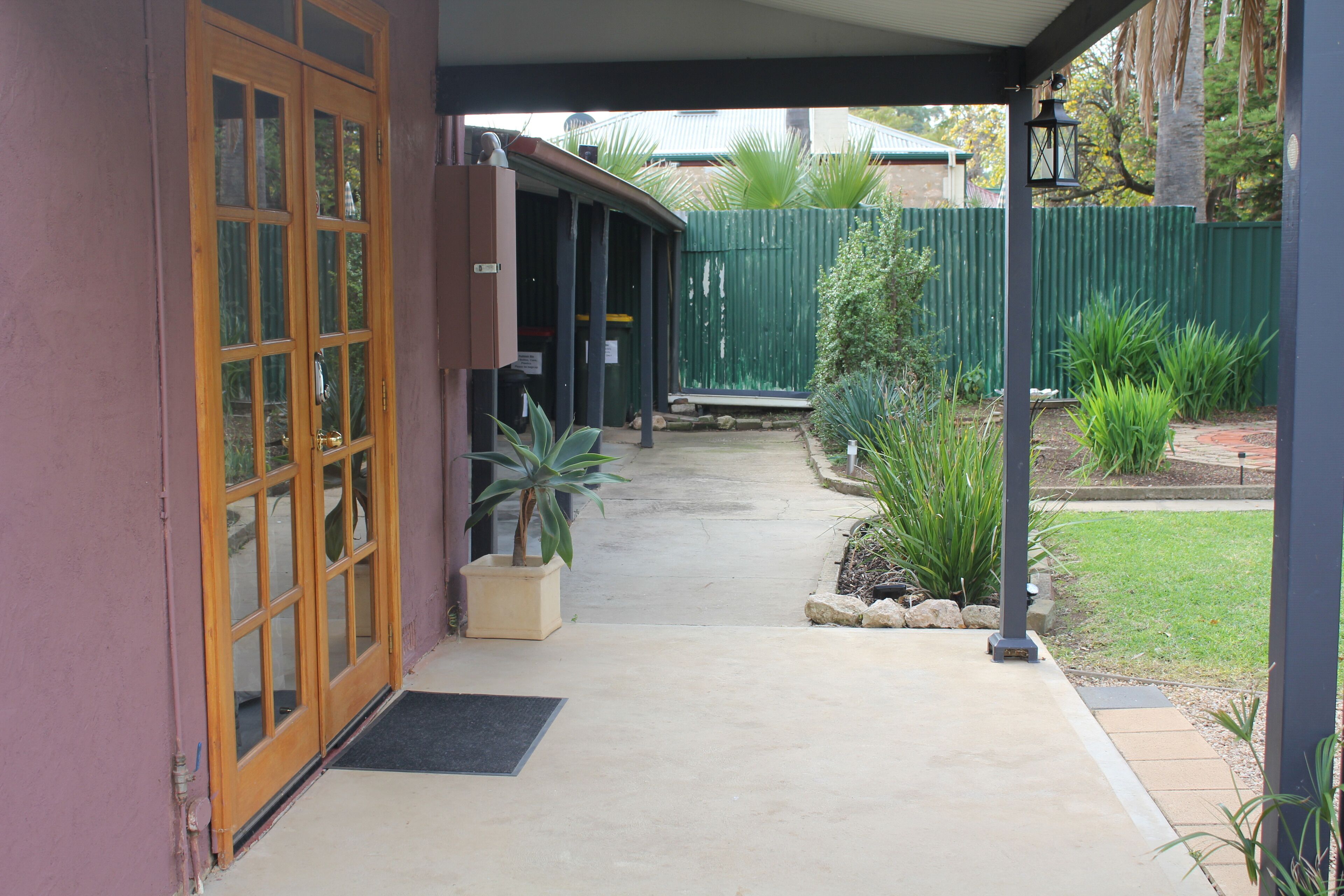 A covered outdoor area with a concrete path leading to a wooden door. There are potted plants and a green fence in the background.