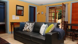 A living room with a black leather sofa, a wooden coffee table, and a dining table with chairs. There is a clock on the wall and a door leading outside. - Unique Cottages Gawler (Gawler South)
