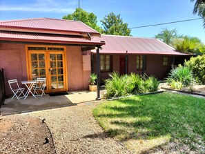 A building with a red roof, a front door, and a small patio area with two white chairs and a table. - Unique Cottages Gawler (Gawler South)