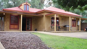 A two-story house with a red roof and beige walls, featuring a patio with outdoor furniture and a barbecue grill. - Unique Cottages Gawler (Gawler South)