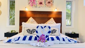 Una cama bien hecha, con una toalla a rayas, una toalla con forma de corazón y un cojín floral. Hay dos mesitas de noche con lámparas y una ventana con vista a palmeras.