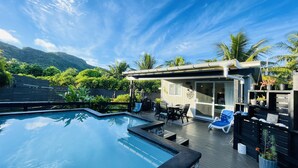 Un área de piscina con terraza de madera, sillas de descanso y una zona de estar cubierta con vistas a las montañas.