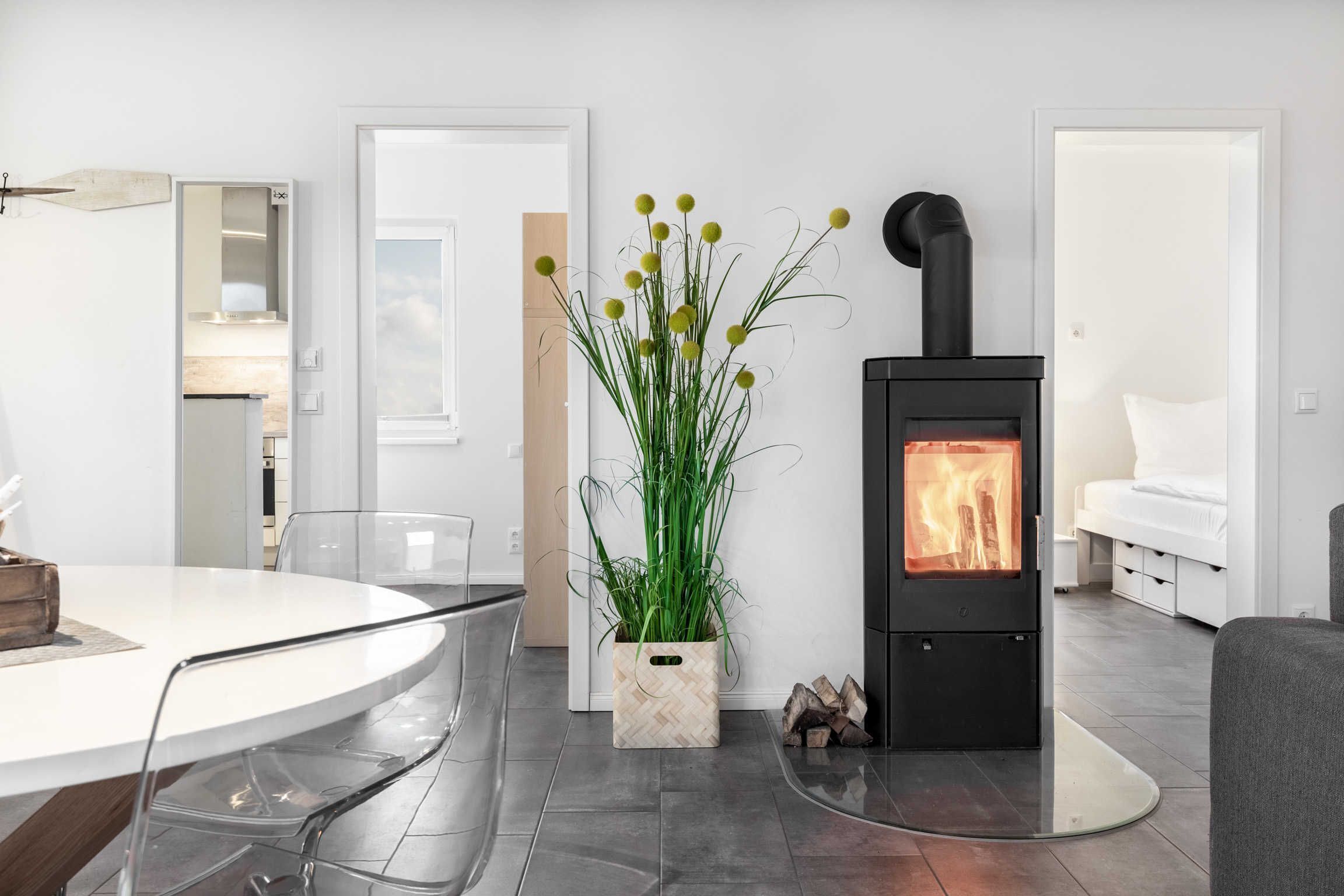 A modern living room with a round glass table, chairs, a potted plant, a black wood-burning stove, and a white sofa.