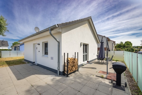 A single-story house with a white exterior, a chimney, a satellite dish, a stack of firewood, and a barbecue grill.