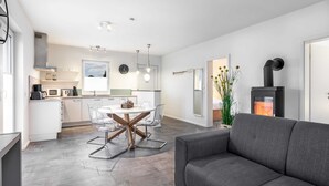 A modern living room with a grey sofa, a dining area with a glass table and chairs, a kitchen with white cabinets, and a black fireplace. - The White BeachHouse - The Blue and The White BeachHouse OFC 16 (Am Salzhaff)
