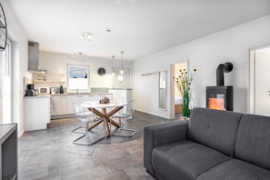 Un salon moderne avec un canapé gris, un coin repas avec une table et des chaises en verre, une cuisine avec des meubles blancs et une cheminée noire.