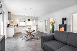 En moderne stue med en grå sofa, et spiseområde med et glasbord og stole, et køkken med hvide skabe og en sort pejs.