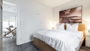 A bedroom with a bed, a nightstand, a wall-mounted lamp, and a painting. - The White BeachHouse - The Blue and The White BeachHouse OFC 16 (Am Salzhaff)