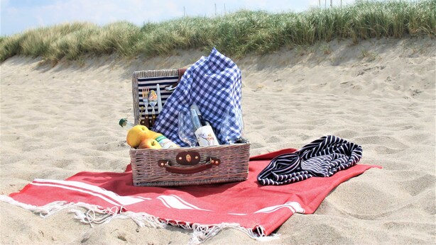A beach scene with a wicker basket containing a bottle, a book, and a fruit on a red towel. - The White BeachHouse - The Blue and The White BeachHouse OFC 16 (Am Salzhaff)
