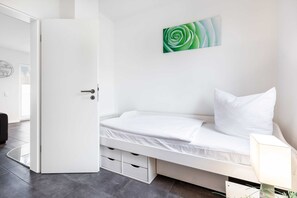 A white room with a bed, a door, a wall-mounted picture, and a small table with a lamp.