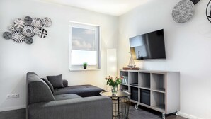 A modern living room with a sectional sofa, a flat-screen TV, a round table with flowers, and a wall-mounted shelf. - The White BeachHouse - The Blue and The White BeachHouse OFC 16 (Am Salzhaff)