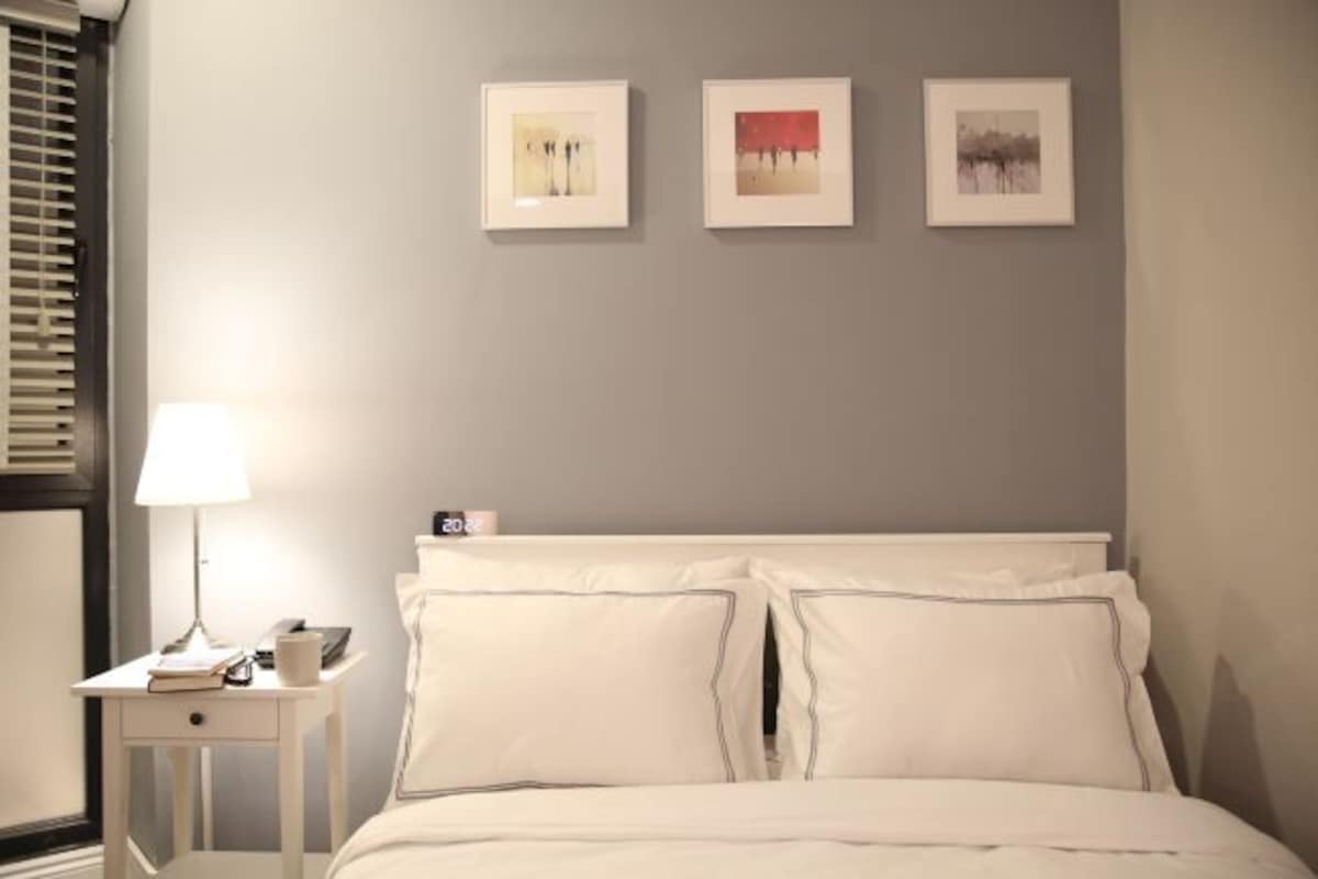 A neatly made bed with white linens, a bedside table with a lamp, and three framed pictures on the wall.