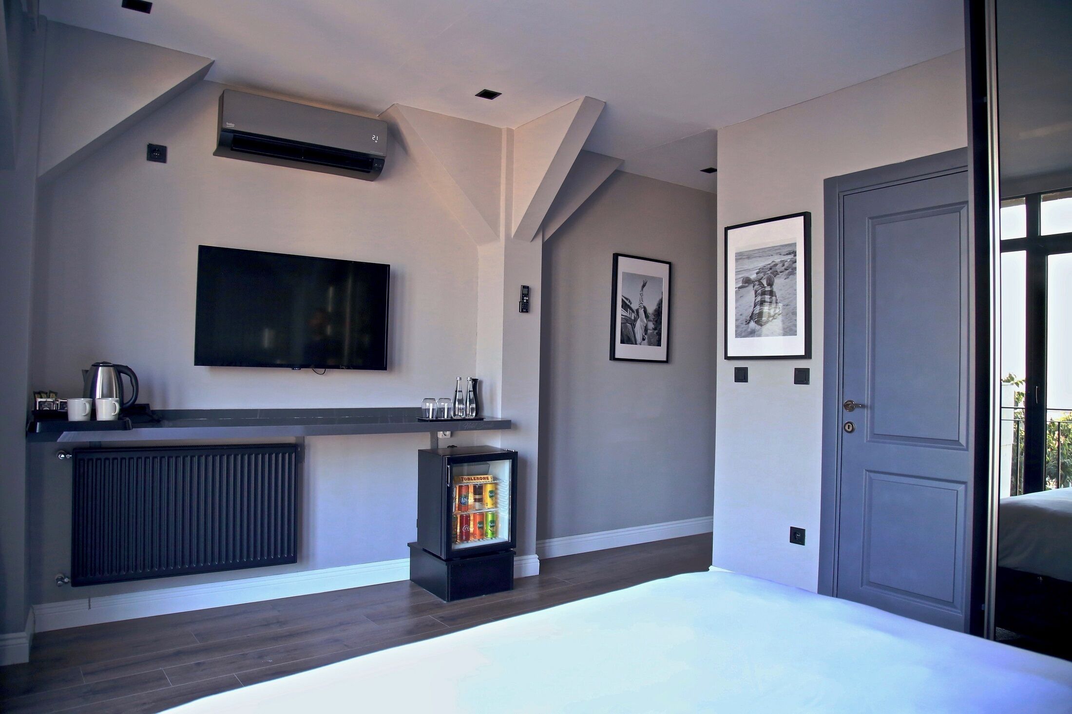 A modern hotel room with a large flat-screen TV, a minibar, a radiator, and a door leading to a balcony.