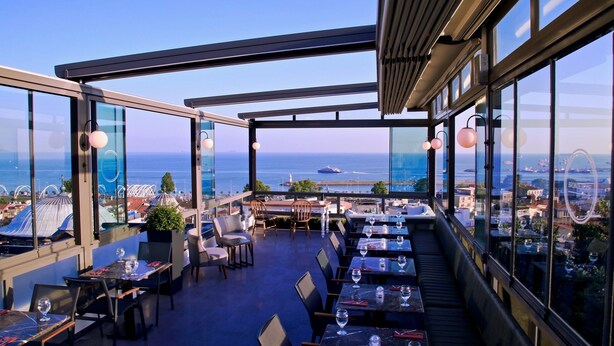 A rooftop restaurant with tables and chairs, large windows offering a view of the sea and cityscape. - Hotel Bronte (Istanbul)