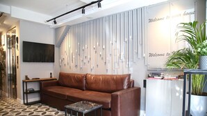 A modern living room with a brown leather sofa, a small black side table, a patterned floor, a wall-mounted TV, a bookshelf, and a decorative plant. - Hotel Bronte (Istanbul)