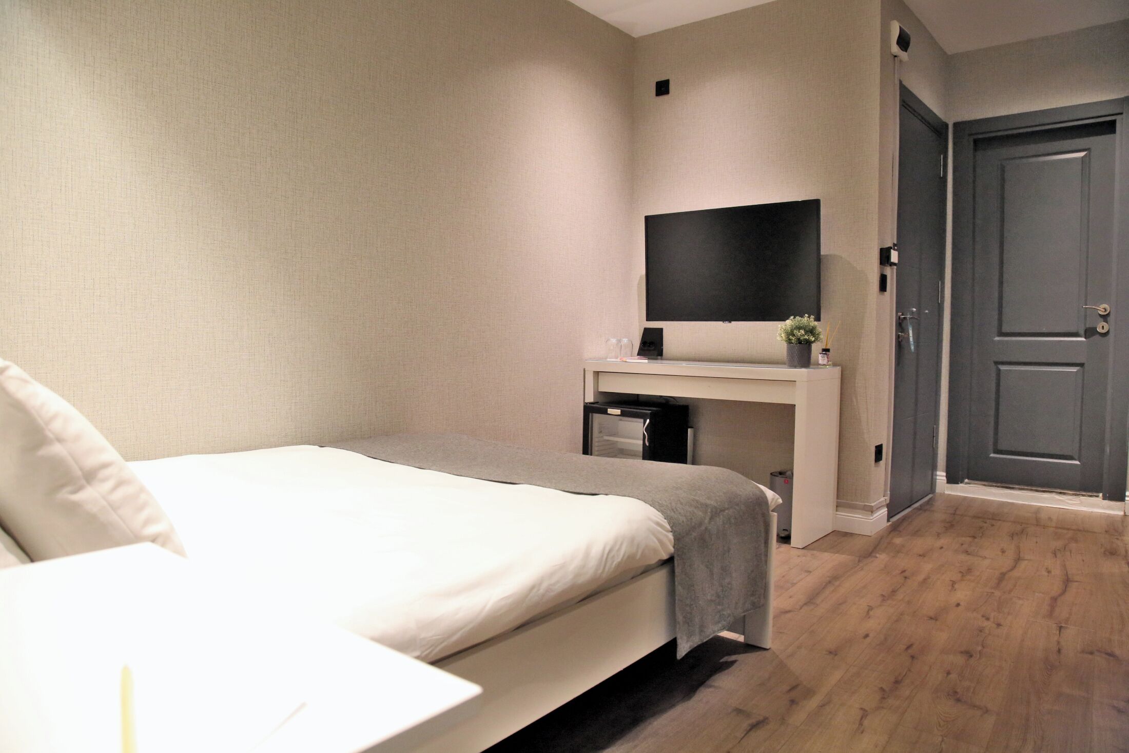 A bedroom with a bed, a desk with a computer, a flat-screen TV, and a door.