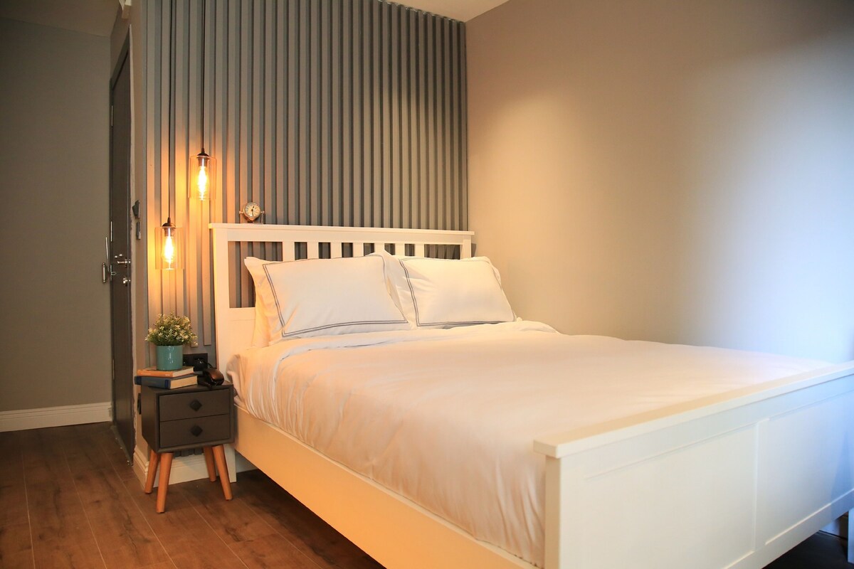 A neatly made bed with white bedding and a wooden headboard, a bedside table with a lamp, and a wall with vertical stripes.