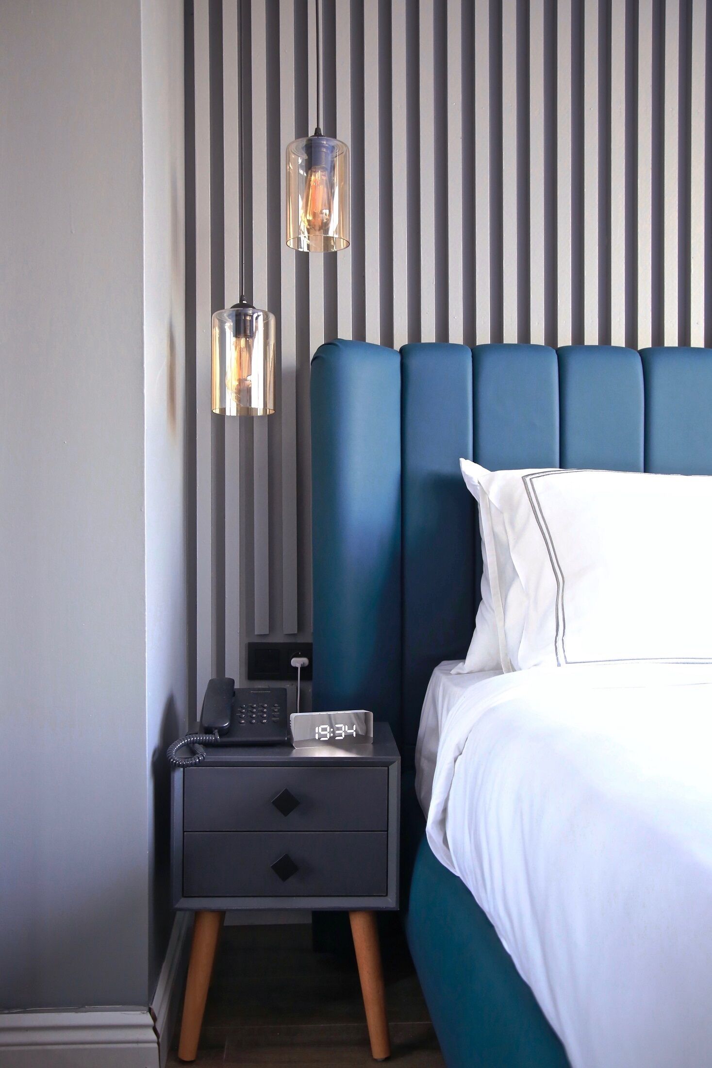 A modern bedroom with a blue upholstered headboard, a white pillow, and a bedside table with a phone and a clock displaying 19:34.