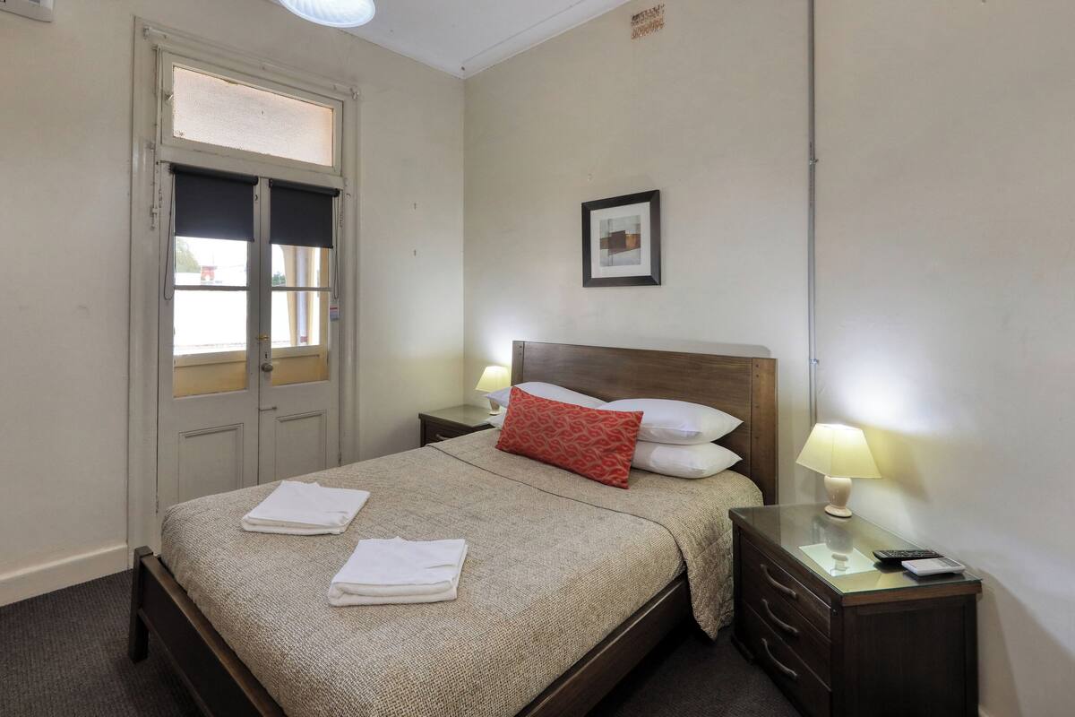 Ein Hotelzimmer mit einem Bett, zwei Nachttischen mit Lampen, einem Holzstuhl und einem Fenster mit Jalousien.