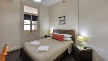 Habitación de hotel con cama, dos mesitas de noche con lámparas, una silla de madera y una ventana con persianas.