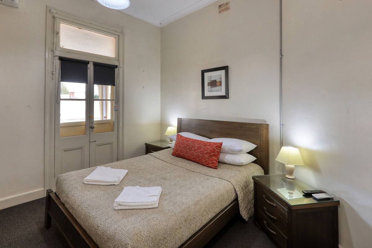 Ein Hotelzimmer mit Bett, zwei Nachttischen, einem Fenster mit Jalousien und einem gerahmten Bild an der Wand.