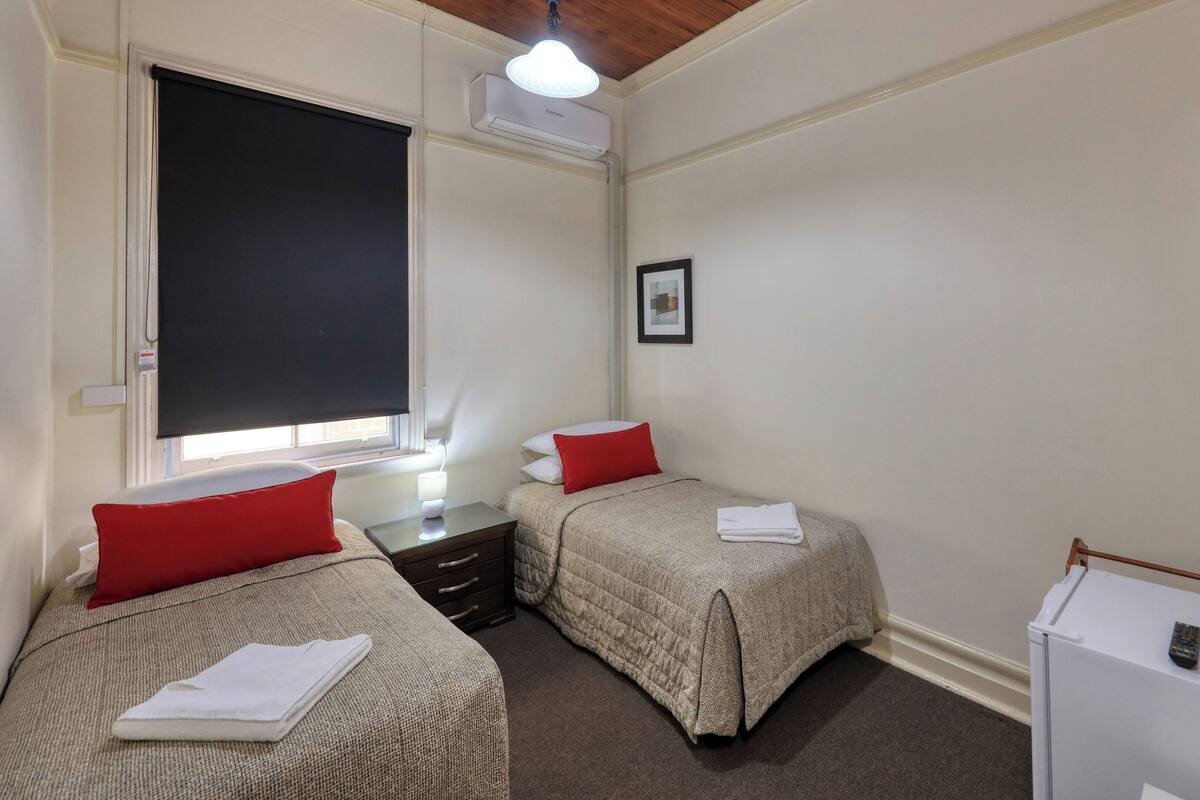 Ein Zimmer mit zwei Betten, jedes mit einem roten Kissen und einem weißen Handtuch, einem kleinen Tisch mit einer Lampe, einem weißen Schrank und einem Fenster mit einem schwarzen Rollo.