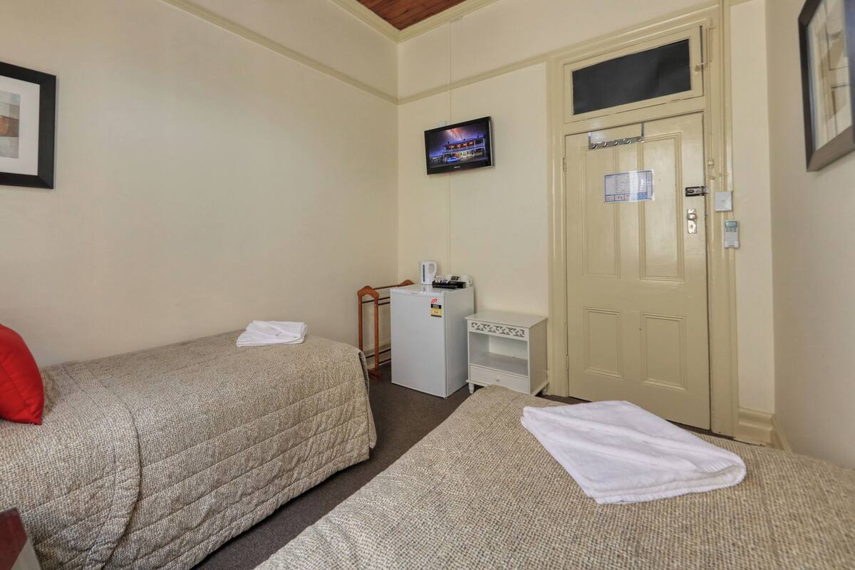 Ein kleines, sauberes Zimmer mit zwei Einzelbetten, einem kleinen Schreibtisch mit Kühlschrank, einem an der Wand montierten Fernseher und einer Tür mit Schloss und Schlüsselöffnung.