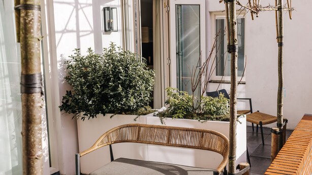 A wicker sofa with a cushion, a small table, and a bench in a sunlit outdoor area with plants and a white wall. - Boutique hotel Margretha (Zandvoort)