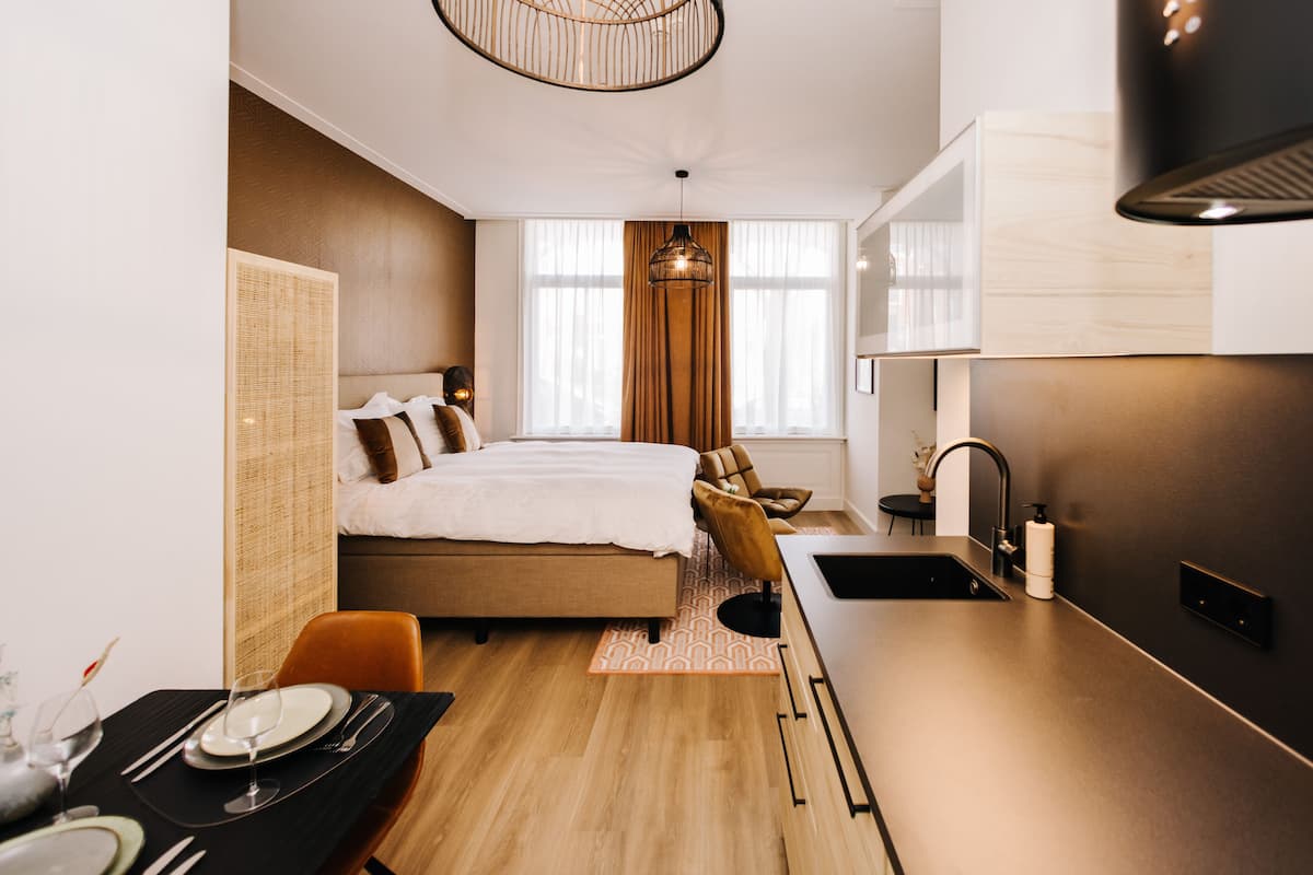 Una habitación de hotel moderna con cama, una mesa de comedor, una zona de cocina y un amplio ventanal con cortinas.