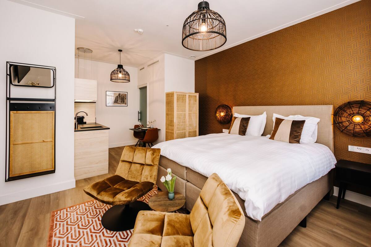 Habitación de hotel moderna con una cama grande, una zona de estar con sofá y sillones, una mesita con un jarrón y una kitchenette.