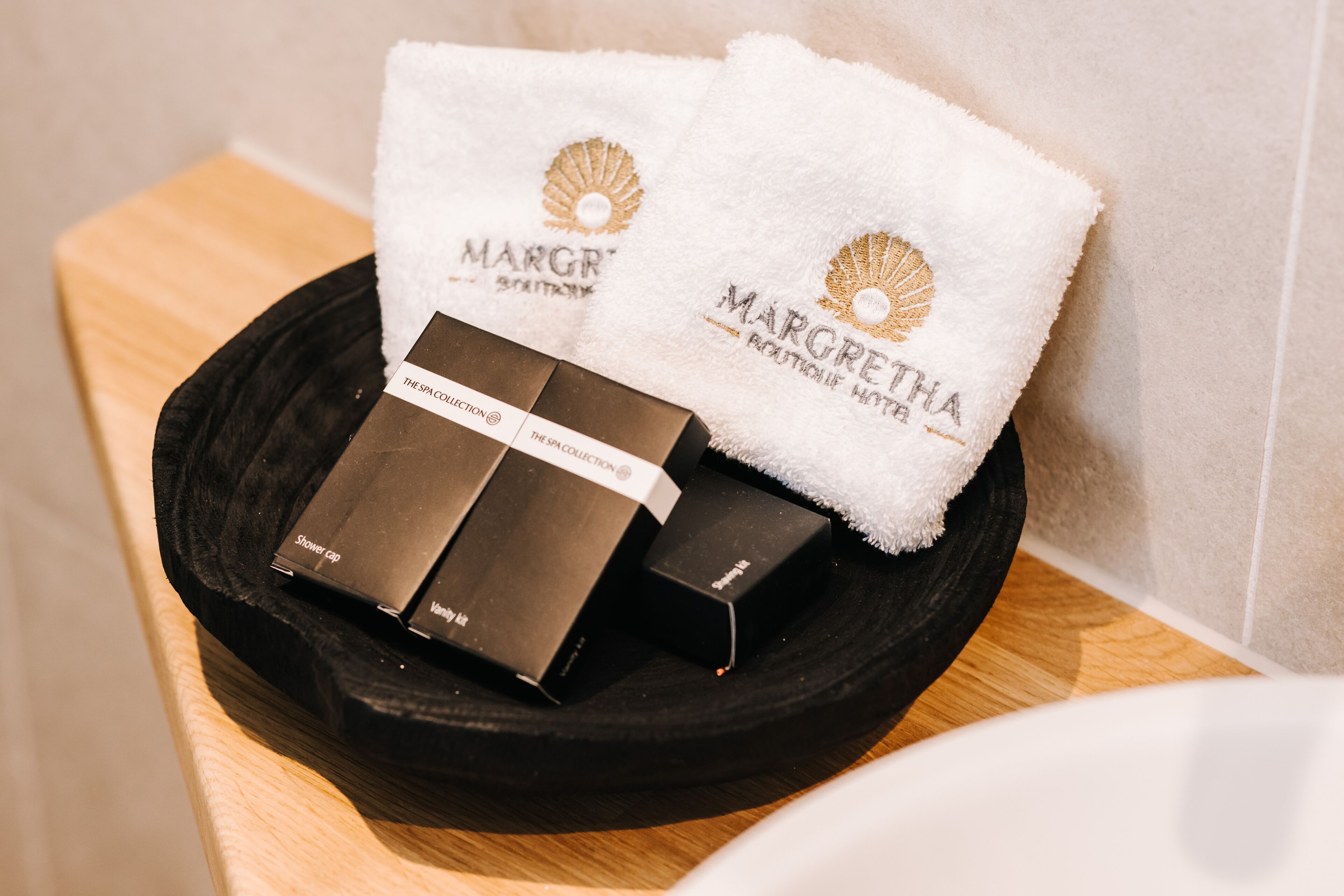 A black tray with three boxes and two white towels with 'MARGRETHA' branding, placed on a wooden surface.