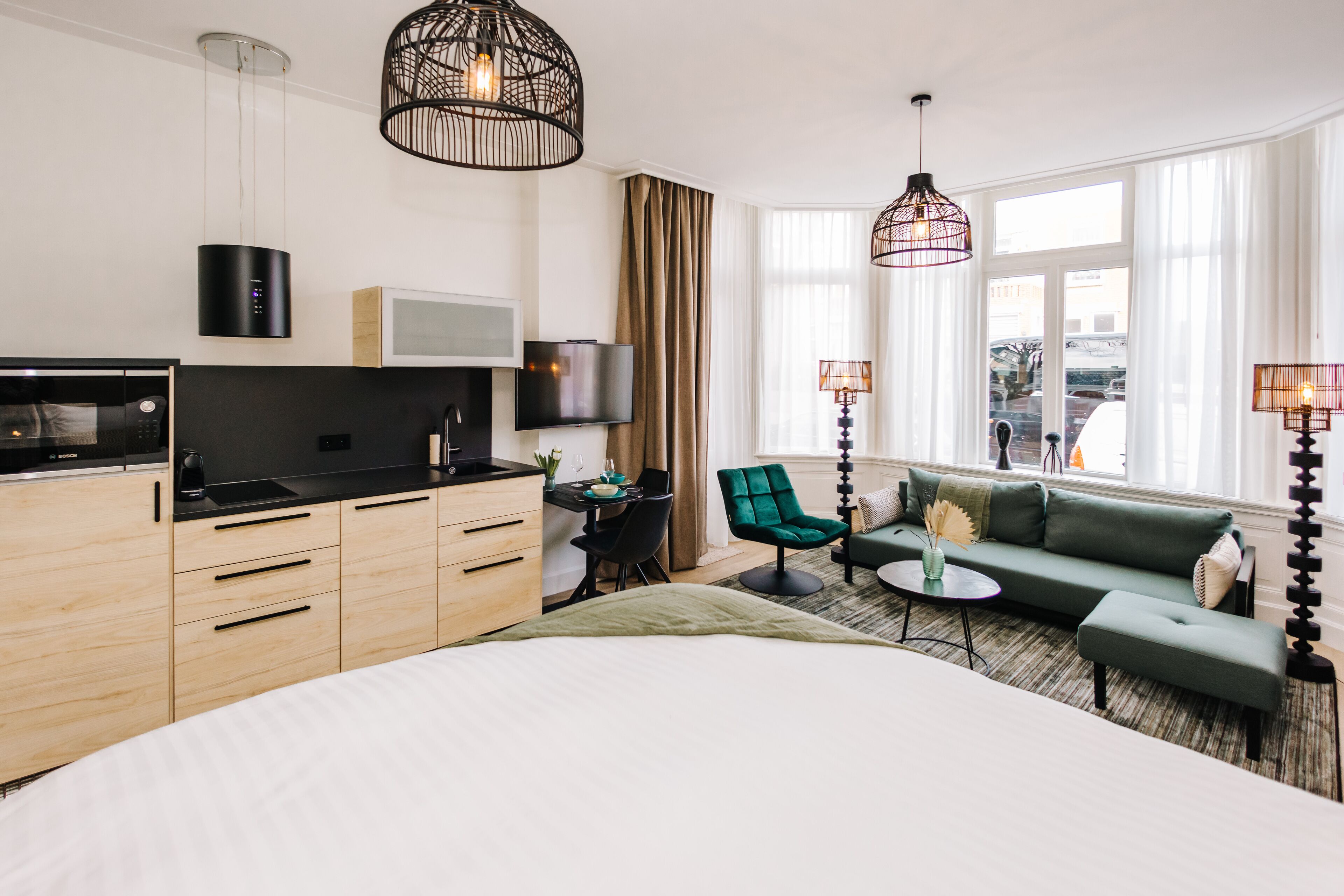 A modern hotel room with a bed, kitchenette, TV, seating area, and large windows.
