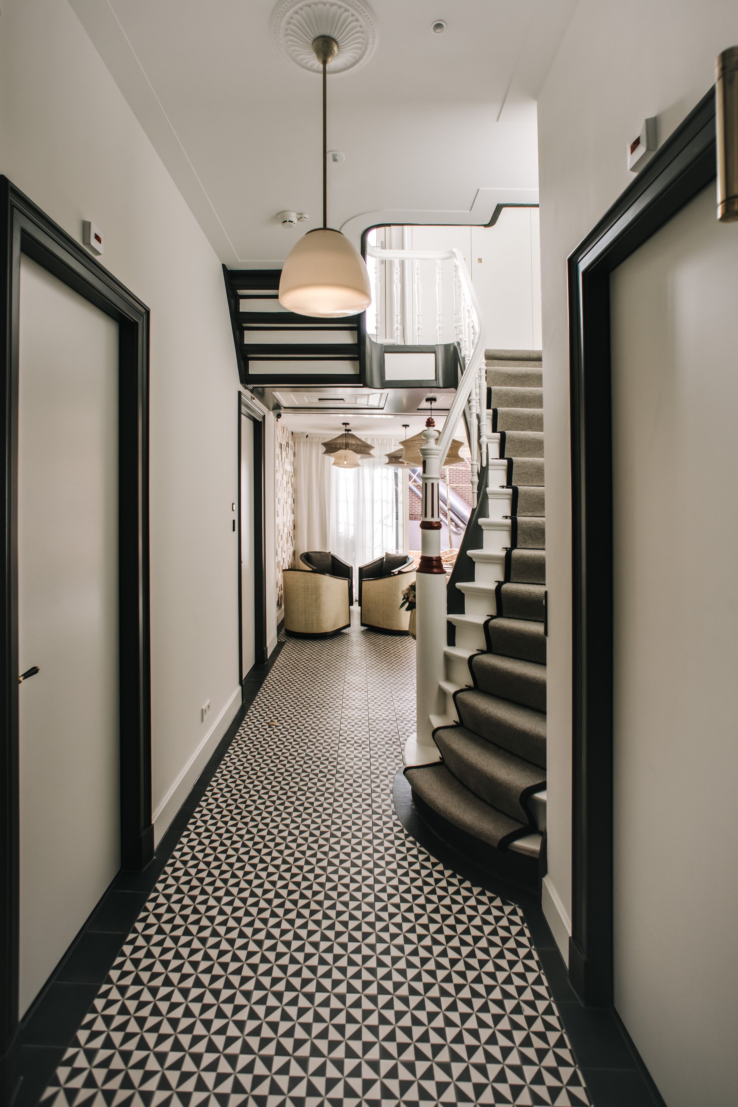 A hallway with a geometric patterned floor, a staircase with a dark carpet, and a pendant light.