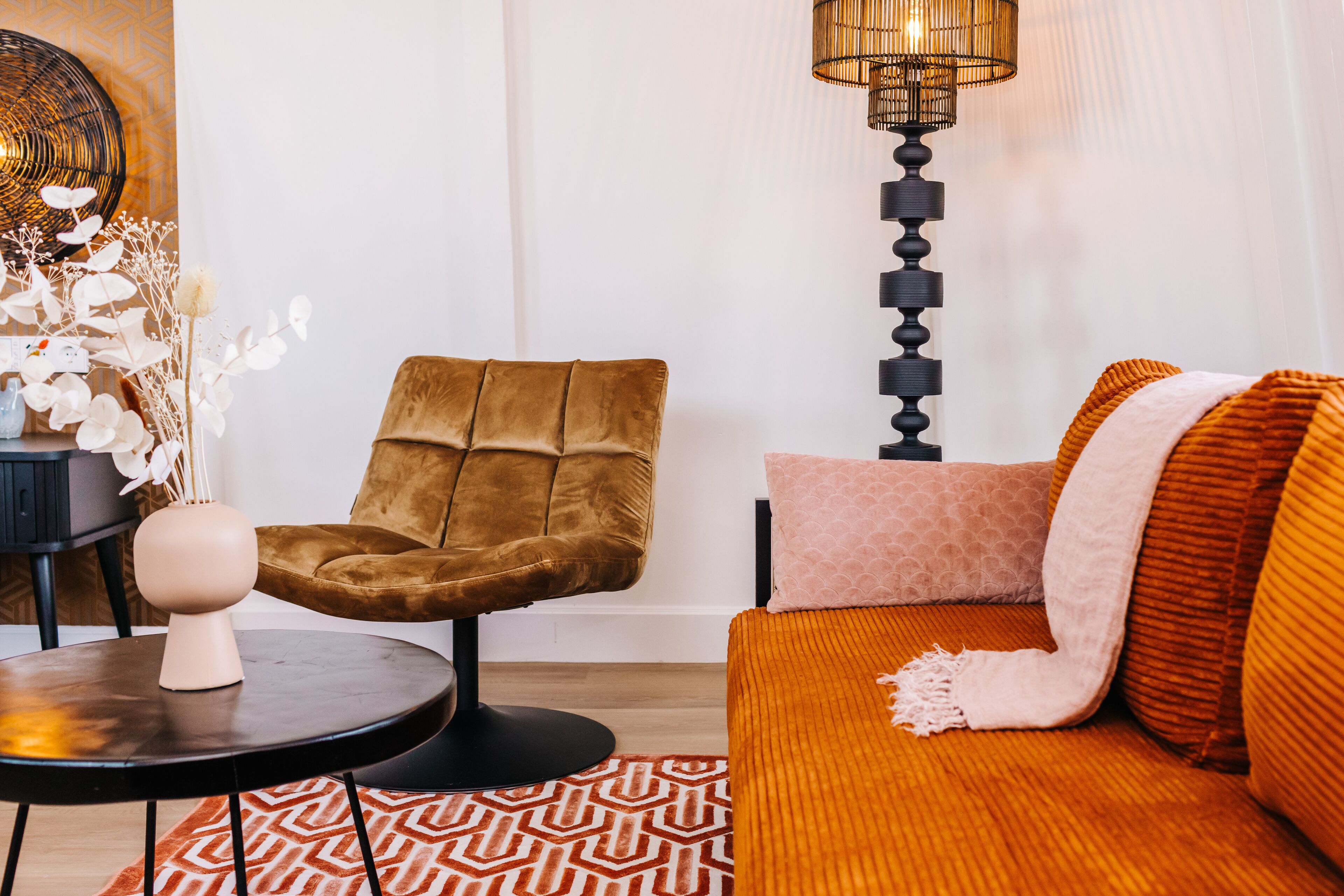 A modern living room with a brown velvet armchair, a patterned rug, and a sofa with orange cushions.