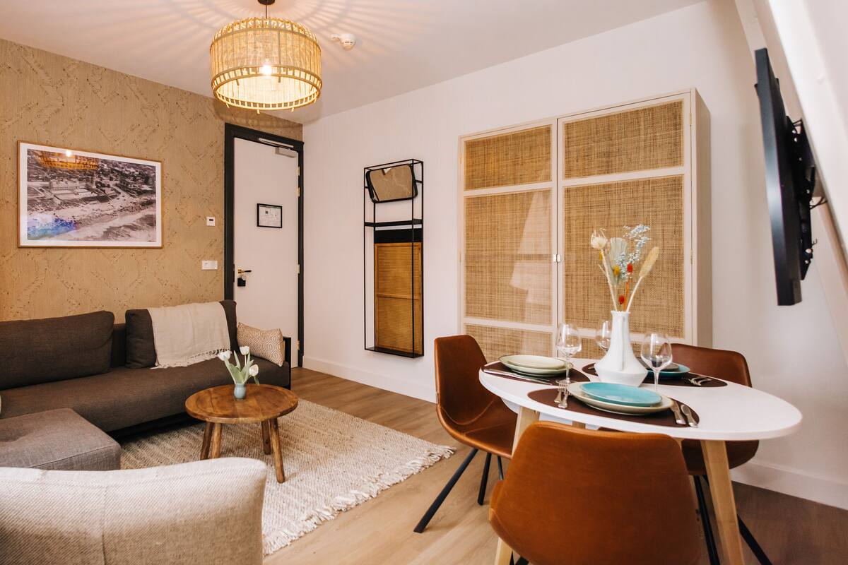 Una sala moderna con un sofá, una mesa de comedor preparada para dos personas y un ventanal con persianas de mimbre.