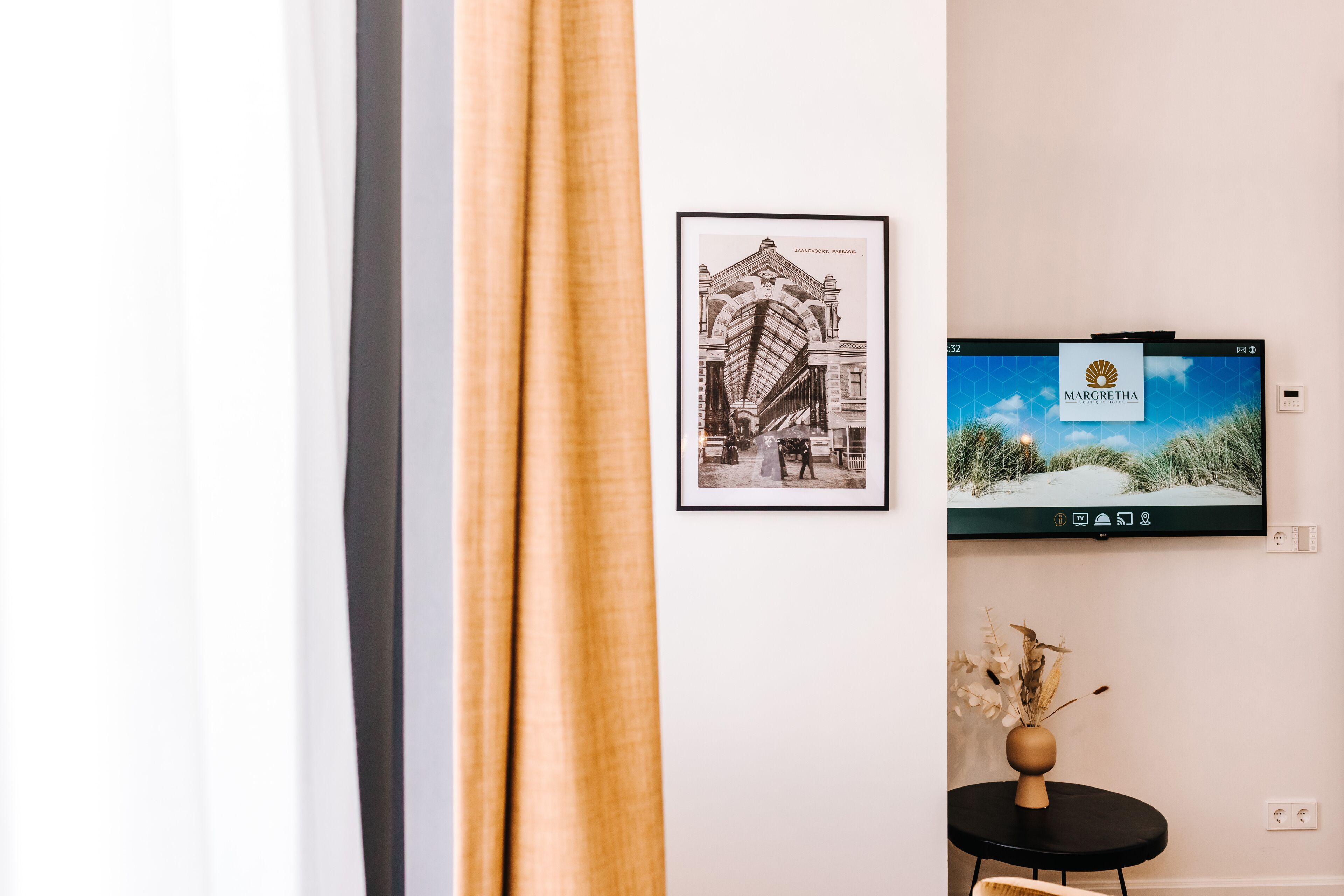 Ein gerahmtes Schwarz-Weiß-Foto von einem Gebäude, einem Fernseher, der das Marge Thoma Logo zeigt, und einem kleinen Tisch mit einer Vase getrockneter Blumen.