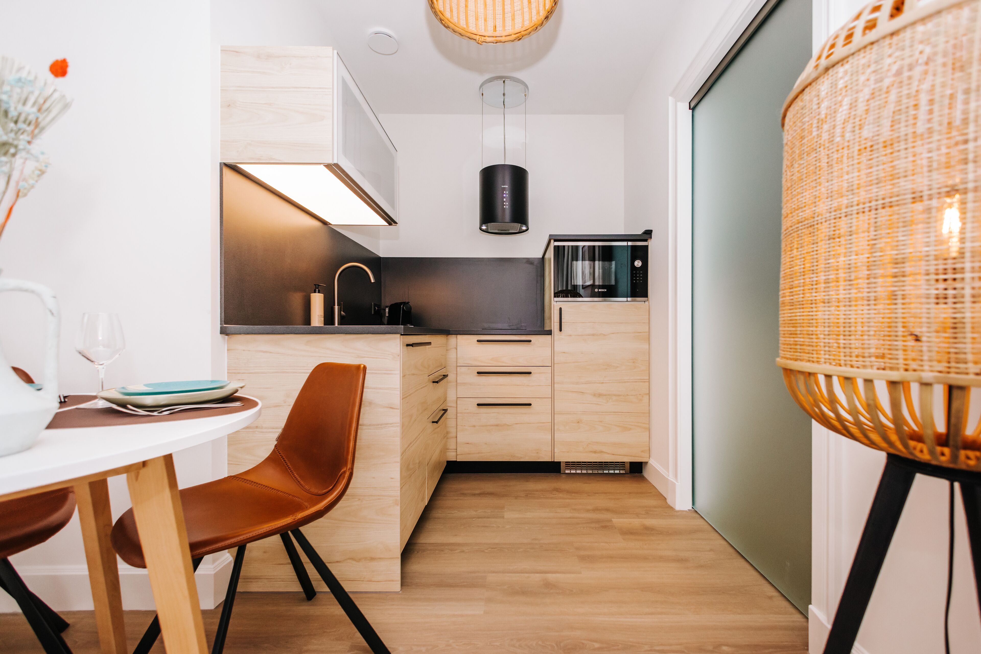 Een compacte keuken met houten kastjes, een ingebouwde magnetron en een hangende lamp.