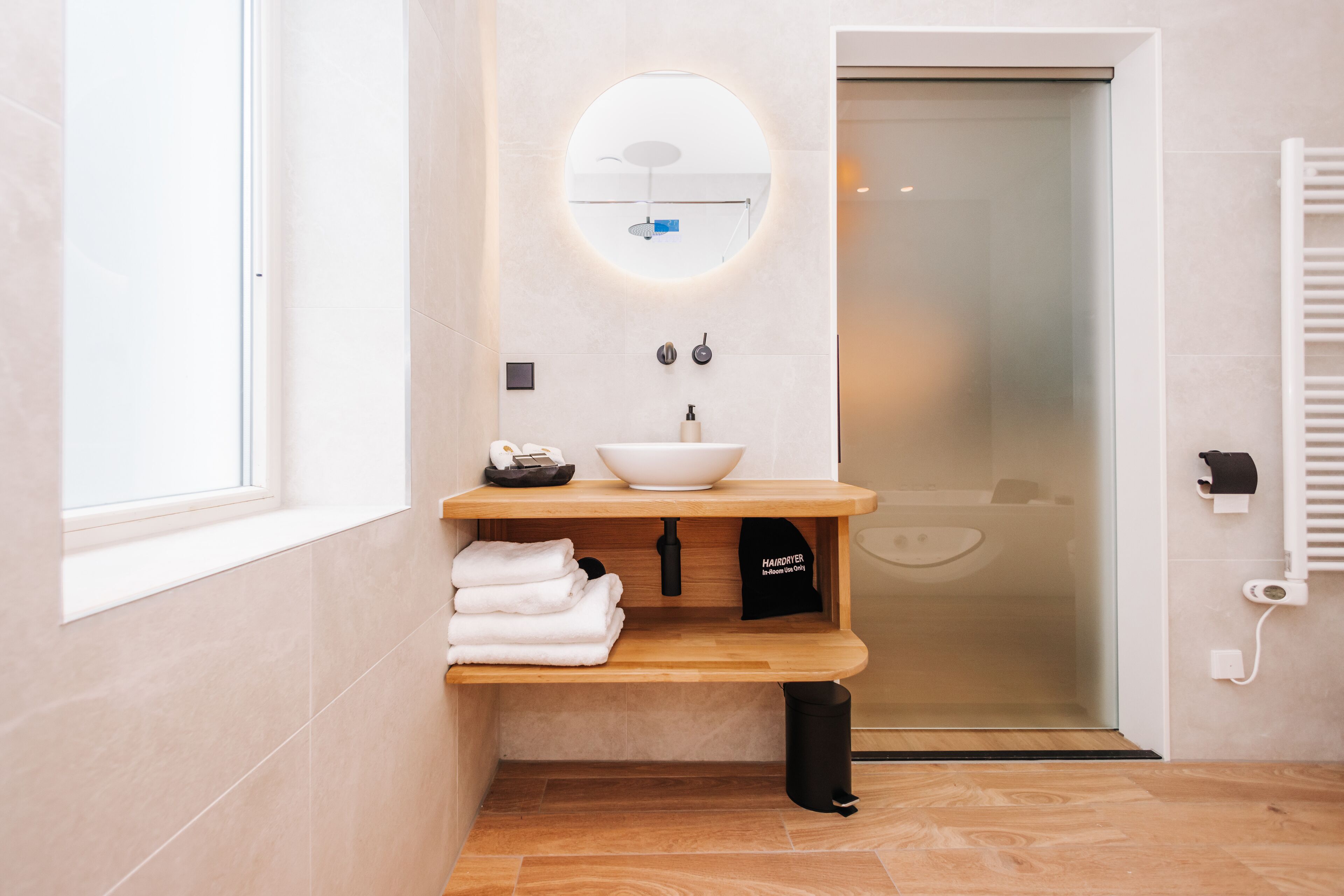 Un baño moderno con tocador de madera, un espejo redondo, un lavamanos blanco y un perchero para toallas.