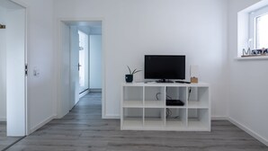 A modern, minimalistic living room with a flat-screen TV on a white entertainment unit, a hallway leading to another room, and a window with a view.