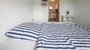 A bedroom with a bed featuring striped bedding, a small bedside table with a potted plant, and a view into the adjacent kitchen area.
