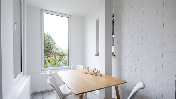 A dining area with a wooden table and white chairs, a white brick wall, and a window with a view of greenery.