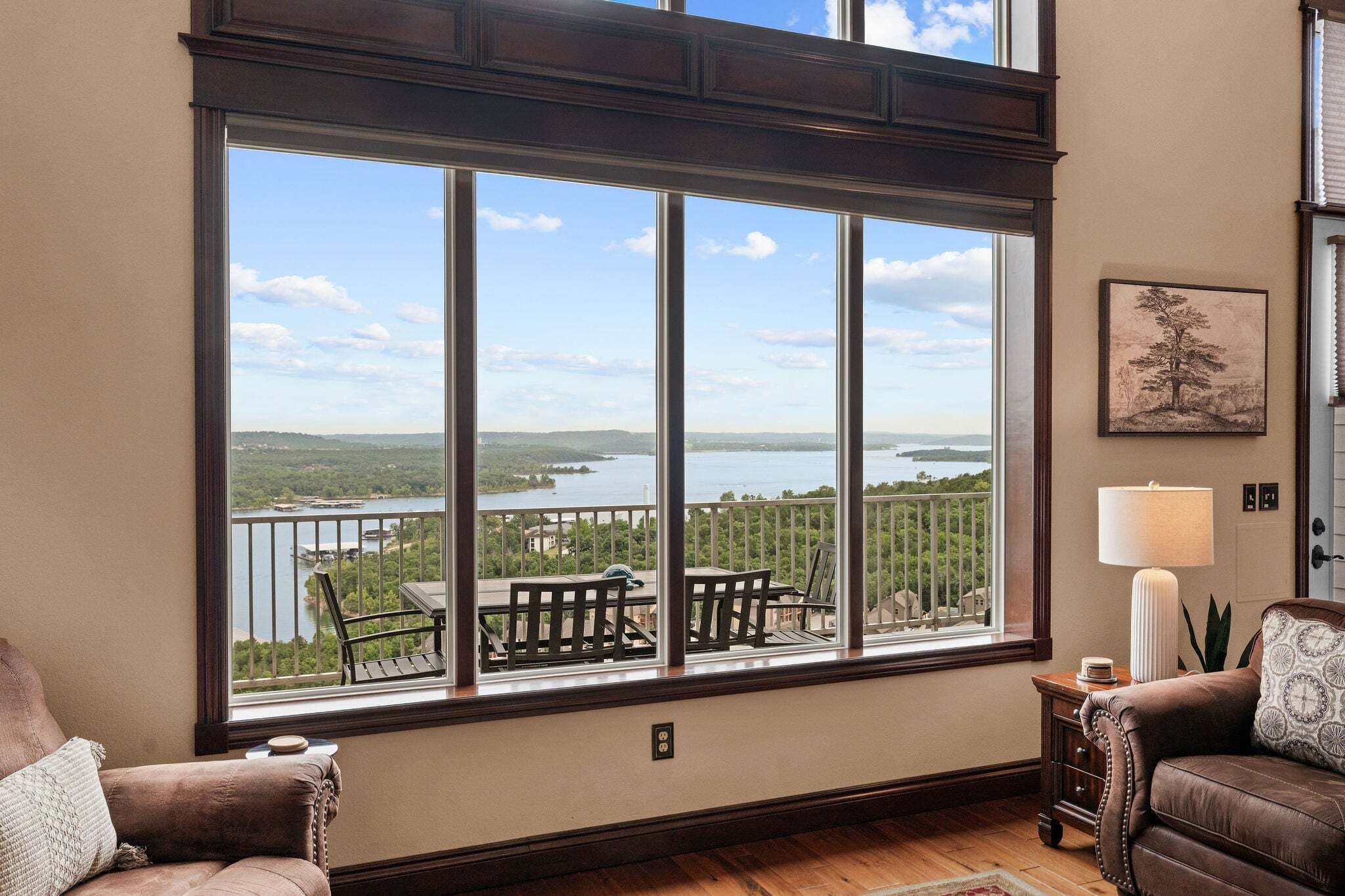 A room with a large window overlooking a lake, a wooden deck with outdoor furniture, and a cozy interior with a sofa and a lamp.
