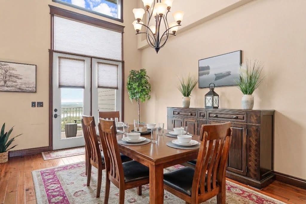 Un coin repas avec une table en bois dressée pour quatre personnes, entourée de chaises. Un lustre est suspendu au-dessus de la table, il y a un meuble contenant des objets décoratifs et une porte qui donne sur un balcon.