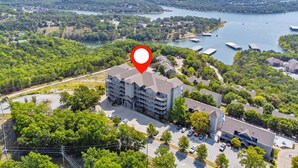 Vista aérea de una zona residencial con un edificio destacado señalado con un alfiler rojo, rodeado de exuberante vegetación y un lago con barcos.