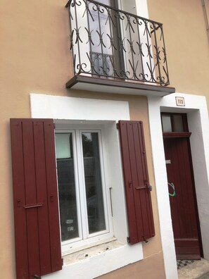 Un edificio con balcón, una ventana con contraventanas rojas y una puerta con placa de número.