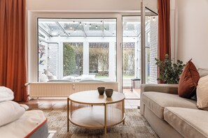 A living room with a sofa, a round table, and a view through a glass door to an outdoor seating area. - Helle und Gemütliche Erdgeschosswohnung in Einem Doppelhaus (Harlesiel)